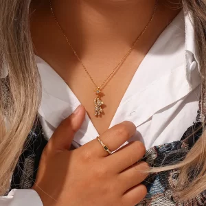 A person wearing a gold teddy bear pendant necklace with cubic zirconia and a matching star charm, paired with a simple gold ring, styled with a white shirt.