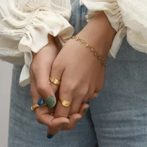 Close-up of hands adorned with an 18K gold-plated heart chain bracelet and statement rings, styled with a ruffled blouse and denim jeans.