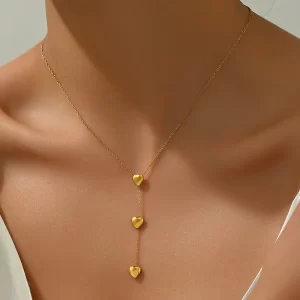 A gold Y-necklace with three small heart-shaped charms, elegantly draping down the neckline of a model wearing a white outfit.