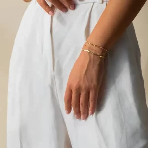 A model wearing a set of thin gold chain bracelets, including herringbone and rope designs, paired with white linen pants for a minimalist aesthetic.