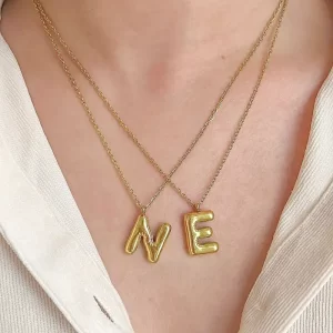 A close-up of a model wearing layered gold necklaces with puffed initial pendants "N" and "E" against a neutral-colored outfit.
