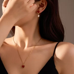 A woman wearing a gold-plated red agate heart necklace and matching pearl drop earrings, styled with a black outfit against a soft-lit background.