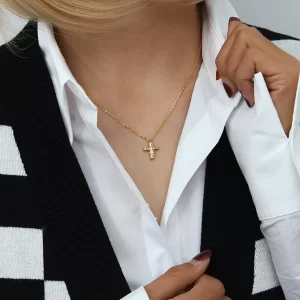 Gold cross necklace with cubic zirconia crystals worn by a person in a white shirt and black cardigan, with a hand adjusting the shirt collar.