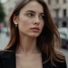 A woman with wavy brown hair wearing 18K gold-plated pearl and heart drop earrings, styled with a black blazer against an urban street backdrop.