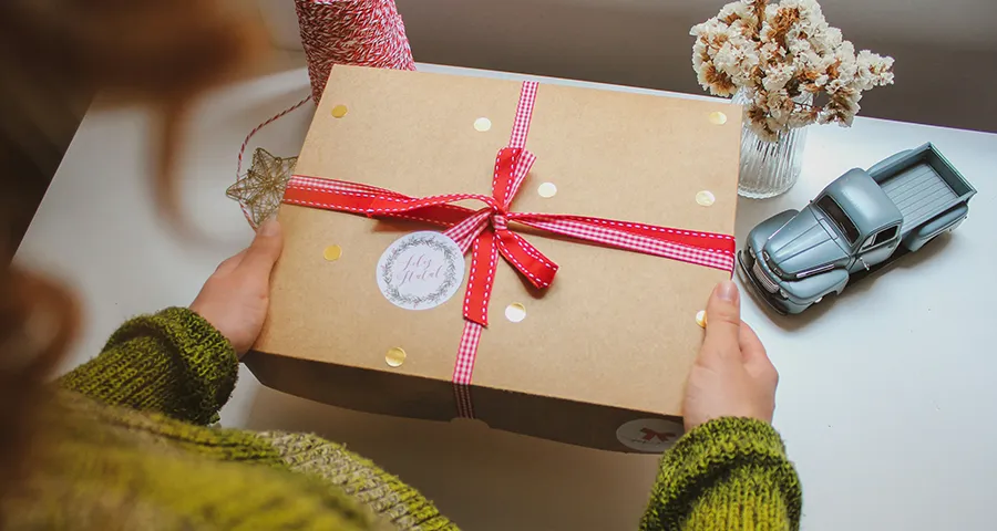 Hands holding a kraft gift box with red ribbon, surrounded by dried flowers and a mini vintage truck in a festive setting